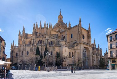 Segovia's massive Cathedral of Santa Mara dominates the plaza. Its Gothic style towers over the bustling public space, where Segovian life unfolds under a blue sky
