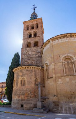 The Romanesque bell tower of San Esteban stands next to its stone apse in Segovia. The multi-tiered arcades are framed by a cypress tree and blue sky