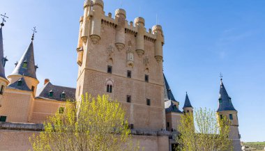 Segovia's famous Alczar stands tall under a blue sky. The fairytale castle features distinctive conical towers, a high keep, and ornate black lampposts lining the path