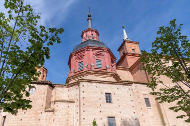 Çarpıcı kırmızı Barok kubbesi ve tarihi tuğla bir kilisenin yüksek kulesi modern bir oyun alanının üzerinde duruyor ve İspanya 'nın Alcal de Henares şehrinde asfalt bir meydan.