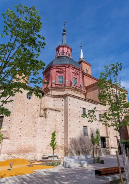 Çarpıcı kırmızı Barok kubbesi ve tarihi tuğla bir kilisenin yüksek kulesi modern bir oyun alanının üzerinde duruyor ve İspanya 'nın Alcal de Henares şehrinde asfalt bir meydan.