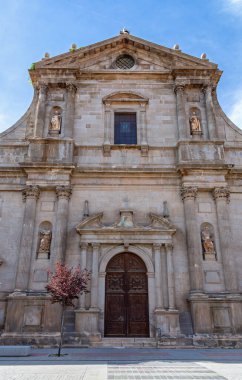 Alcal de Henares 'deki San Ildefonso Kilisesi' nin heykel, sütunlar ve mavi gökyüzünün altında büyük, koyu ahşap bir ana kapı yer alıyor..