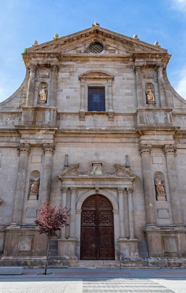 Alcal de Henares 'deki San Ildefonso Kilisesi' nin heykel, sütunlar ve mavi gökyüzünün altında büyük, koyu ahşap bir ana kapı yer alıyor..