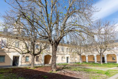 Madrid, El Pardo 'da sakin bir konut avlusu, açık sarı cepheli kemerli bir kapı, çıplak dallı hareketsiz ağaçlar, ve karışık çimenler ve toprak zemin