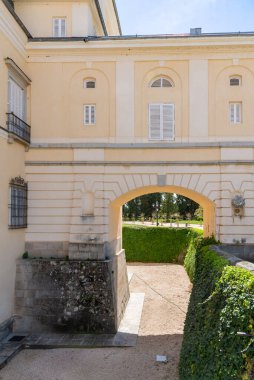 El Pardo Sarayı 'nın soluk sarı duvarı çakıllı bir geçide uzanan taş kemerli uzun, canlı yeşil bir sarmaşık çiti ve sarayın taş temeli ile kaplı.