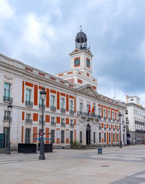 Madrid 'in Puerta del Sol kentindeki tarihi Real Casa de Correos' ta, ikonik saat kulesi, kırmızı tuğla ve beyaz taş cephe ve merkez balkondan dalgalanan İspanyol ve AB bayrakları yer alıyor.