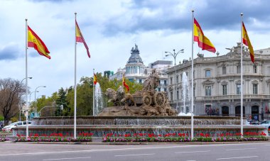 The Fuente de Cibeles in Madrid, featuring Cybele on a chariot pulled by lions, is surrounded by Spanish flags, a flower bed, and a fountain pool under a cloudy sky