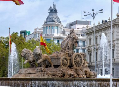 The Fuente de Cibeles in Madrid, featuring Cybele on a chariot pulled by lions, is surrounded by Spanish flags, a flower bed, and a fountain pool under a cloudy sky