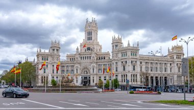 The elegant white Palacio de Cibeles in Madrid, with its towers and detailed stonework, rises over a busy street corner beneath a cloudy, dramatic sky in muted light