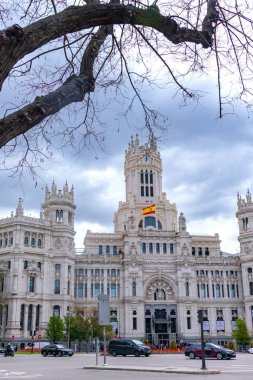 The elegant white Palacio de Cibeles in Madrid, with its towers and detailed stonework, rises over a busy street corner beneath a cloudy, dramatic sky in muted light