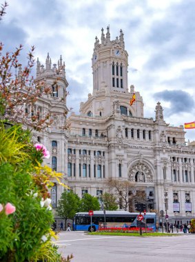 The elegant white Palacio de Cibeles in Madrid, with its towers and detailed stonework, rises over a busy street corner beneath a cloudy, dramatic sky in muted light