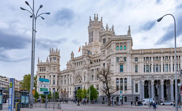 The elegant white Palacio de Cibeles in Madrid, with its towers and detailed stonework, rises over a busy street corner beneath a cloudy, dramatic sky in muted light