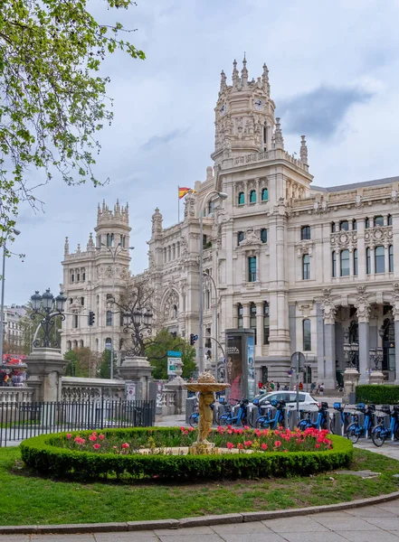 The elegant white Palacio de Cibeles in Madrid, with its towers and detailed stonework, rises over a busy street corner beneath a cloudy, dramatic sky in muted light