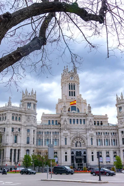The elegant white Palacio de Cibeles in Madrid, with its towers and detailed stonework, rises over a busy street corner beneath a cloudy, dramatic sky in muted light