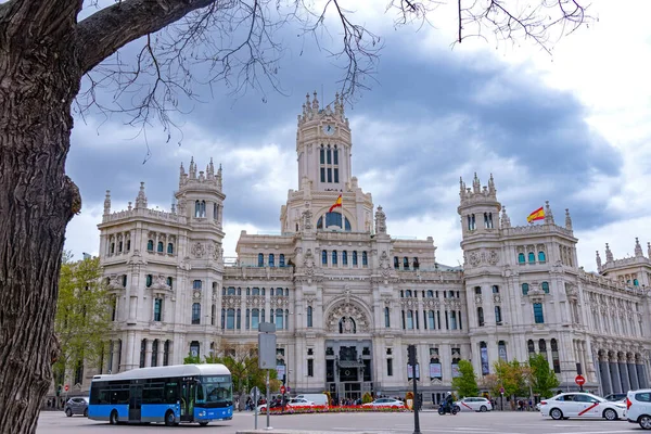 The elegant white Palacio de Cibeles in Madrid, with its towers and detailed stonework, rises over a busy street corner beneath a cloudy, dramatic sky in muted light