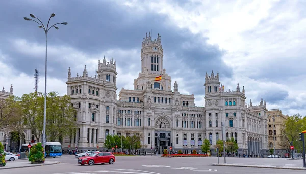The elegant white Palacio de Cibeles in Madrid, with its towers and detailed stonework, rises over a busy street corner beneath a cloudy, dramatic sky in muted light