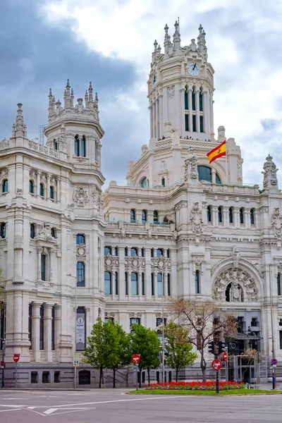 The elegant white Palacio de Cibeles in Madrid, with its towers and detailed stonework, rises over a busy street corner beneath a cloudy, dramatic sky in muted light