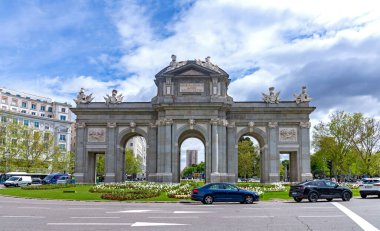 The Puerta de Alcal, a large neoclassical stone triumphal arch in Madrid, features three main arches, detailed carvings, and statues on the roofline under a dramatic, cloudy sky