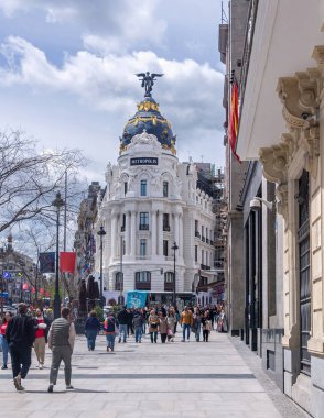 A busy Madrid sidewalk with pedestrians leads toward the Metropolis Building, showing its white facade, dark dome with gold accents, and winged figure under cloudy skies