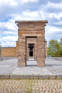 Madrid 'deki Debod Tapınağı, eski bir Mısır anıtı, iki yıpranmış taş sütun ve bir taş plazanın üzerine kurulmuş, ağaçlardan oluşan bir arka plan ve bulutlu mavi gökyüzü.