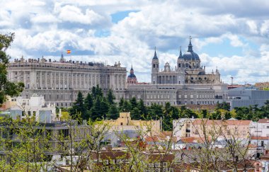 A panoramic view of the Madrid skyline featuring the Royal Palace on the left and the Almudena Cathedral domes on the right, rising above a foreground of lush trees and residential rooftops