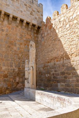 Bir sütunu tasvir eden modern, soyut bir taş heykel ve iki figür, taşın tabanına kırmızı yazılar çizilmiş, büyük, kaba yontulmuş ortaçağ vila duvarlarının gölgesinde duruyor.