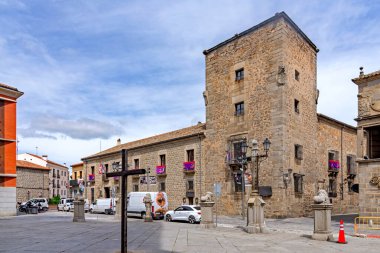 Kare kulesi ve küçük balkonları olan tarihi taş bir saray villadaki katedralin karşısında duruyor, ahşap bir haçı ve park edilmiş araçları olan bir plazaya bakıyor.