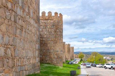Taştan kuleleri olan vila duvarları ve park halindeki arabalarla dolu bir yolun yanında çimenli bir yamaç, yukarıdaki yumuşak mavi gökyüzünün altındaki uzak manzarayla çevrelenmiş.