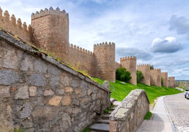 Villa duvarlarından bir sıra taş kule yükselir dik, parlak yeşil bir setin üzerinde, ön planda alçak bir taş duvar, adımlar ve küçük bahar çiçekleri ile çerçevelenmiş.