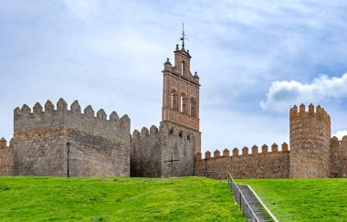 Yüksek, ince, kırmızımsı kahverengi taş bir şapel ya da istihkamın içine inşa edilmiş bir gözetleme kulesinin yer aldığı dik yeşil bir tepenin bulunduğu vila duvarlarının manzarası.