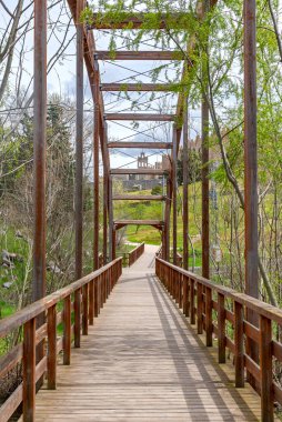 Uzun ahşap bir yaya köprüsü ve iskeleti, gözü ağaçlık bir parka, uzak bir taş gözetleme kulesine ve dik yeşil bir yamaçtaki alçak inşa edilmiş vila duvarlarına götürüyor.