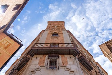 Toledo 'da yüksek desenli tuğla ve taş köşeli bir binanın dinamik manzarası, dövmeli demir bir balkonu ve parlak, bulutlu bir gökyüzüne karşı yükselen kare bir kulesi var.