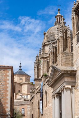 Toledo Katedralleri 'nin yakın çekimleri kubbeyi süslüyor ve parlak mavi gökyüzüne karşı kule yapıyor. Gökyüzünde gotik kemerler ve tepeler bir cadde üzerinde yükseliyor ve arkasında daha küçük bir kubbe var.