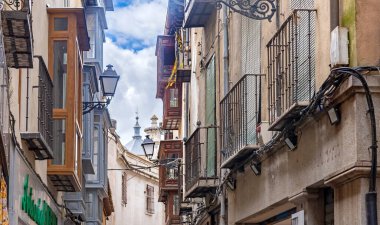 Tahta koy pencereleri ve işlenmiş demir balkonlarla tanımlanan çok dar bir Toledo caddesine yakın plan; uzaklıktan tarihi bir kule görünüyor