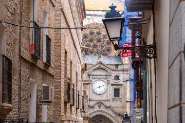 Tahta koy pencereleri ve işlenmiş demir balkonlarla tanımlanan çok dar bir Toledo caddesine yakın plan; uzaklıktan tarihi bir kule görünüyor