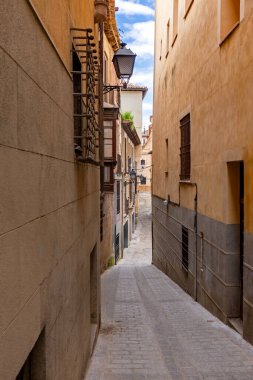 Toledo 'da dar, dolambaçlı bir kaldırım taşından aşağı dikey bir manzara. Bulutlu bir gökyüzünün altında dövülmüş demir balkonları olan tarihi binalar tarafından çerçevelenmiş.