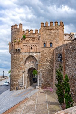 Toledo 'daki Puerta del Cambrn, at nalı kemerli Mudjar taşından bir kapı, yarılmış kuleler ve oyulmuş kabartmalar, bulutlu bir gökyüzünün altındaki şehir duvarları içinde geçiyor.