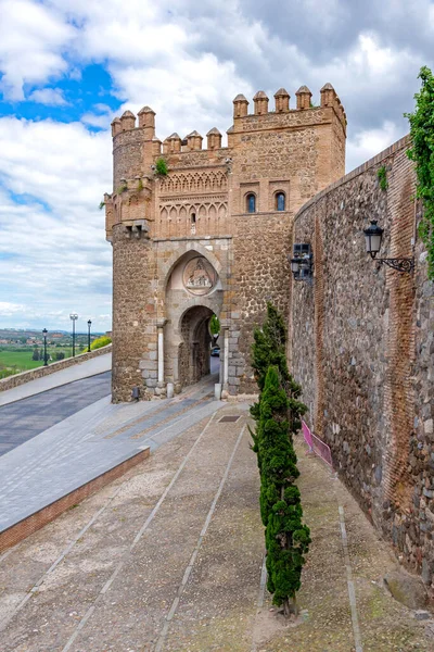 Toledo 'daki Puerta del Cambrn, at nalı kemerli Mudjar taşından bir kapı, yarılmış kuleler ve oyulmuş kabartmalar, bulutlu bir gökyüzünün altındaki şehir duvarları içinde geçiyor.