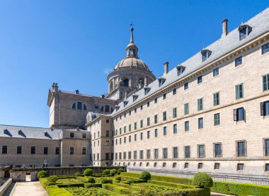 İspanya 'daki El Escorial' ın yan görüntüsü, geometrik boxwood bahçelerini, kayrak tavanlı uzun granit cepheyi ve mavi gökyüzünün altındaki merkezi bazilika kubbesini gösteriyor.