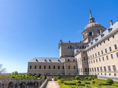 İspanya 'daki El Escorial' ın yan görüntüsü, geometrik boxwood bahçelerini, kayrak tavanlı uzun granit cepheyi ve mavi gökyüzünün altındaki merkezi bazilika kubbesini gösteriyor.