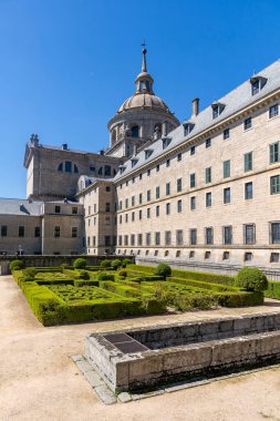İspanya 'daki El Escorial' ın yan görüntüsü, geometrik boxwood bahçelerini, kayrak tavanlı uzun granit cepheyi ve mavi gökyüzünün altındaki merkezi bazilika kubbesini gösteriyor.
