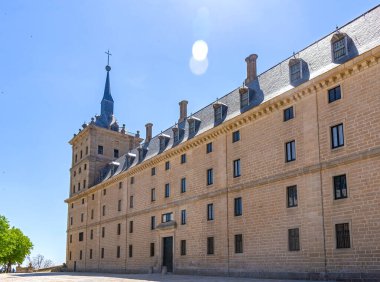 San Lorenzo 'daki ana giriş avlusundan El Escorial Manastırı' nın uzun granit cephesinin güneşli manzarası, kayrak tavanlar ve parlak mavi bir gökyüzü.