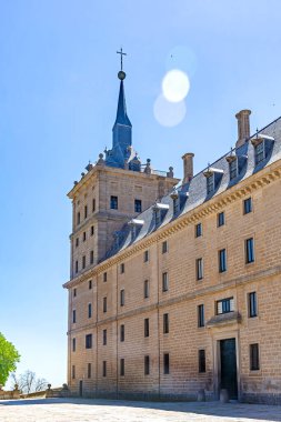 San Lorenzo 'daki ana giriş avlusundan El Escorial Manastırı' nın uzun granit cephesinin güneşli manzarası, kayrak tavanlar ve parlak mavi bir gökyüzü.