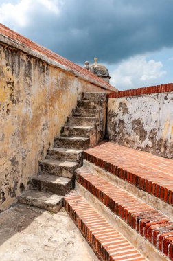 Yıpranmış bir taş merdiven ve kırmızı tuğla katlar dramatik ve bulutlu bir tropikal gökyüzü altında Cartagena, Bolvar, Kolombiya 'nın tarihi savunma duvarlarına tırmanıyor.