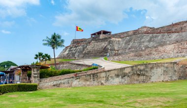 San Felipe de Barajas Kalesi 'nin devasa taş duvarları, Kolombiya' nın başkenti Cartagena 'da bulutlu bir gökyüzünün altında yeşil çimlerin ve palmiye ağaçlarının üzerinde yükselir.
