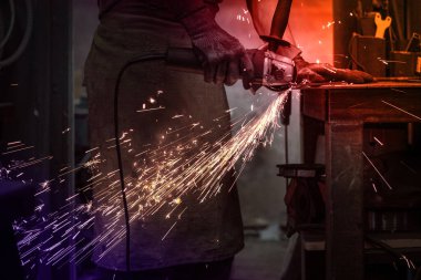 Bir işçi metali işliyor, bir demircinin elinde bir disk elektrik makinesi dairesel bir testereden parlayan kıvılcımlar çıkarıyor.