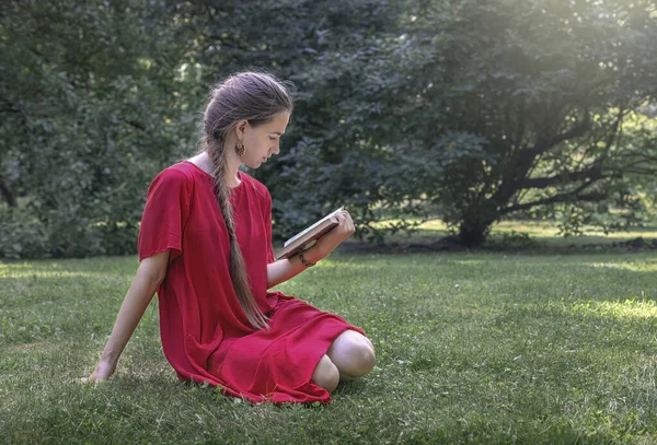 young woman reading book in summer park, a girl in a red dress sitting in the park on the grass holding a book in her hands