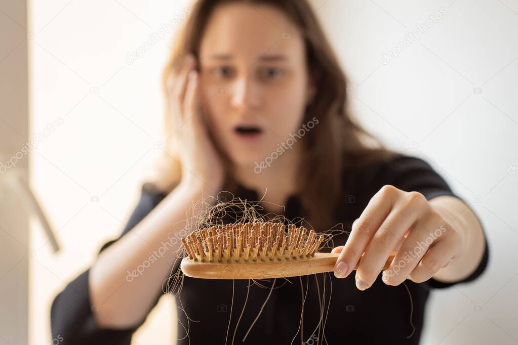 Young woman holding a comb with a lock of hair in front of her, focus on comb, hair loss problem, a lot of long hair on a wooden brush after combing