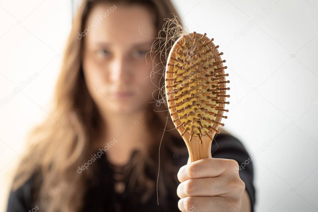 Young woman holding a comb with a lock of hair in front of her, focus on comb, hair loss problem, a lot of long hair on a wooden brush after combing