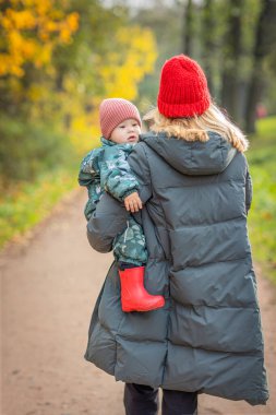 Bir bebekle parkta yürüyüş, anne kucağında bir çocuğu kucağında sokakta, arkadan manzara, kalın elbiseler, parlak şapkalar, yaşam tarzı, yaprak düşüşü sezonu